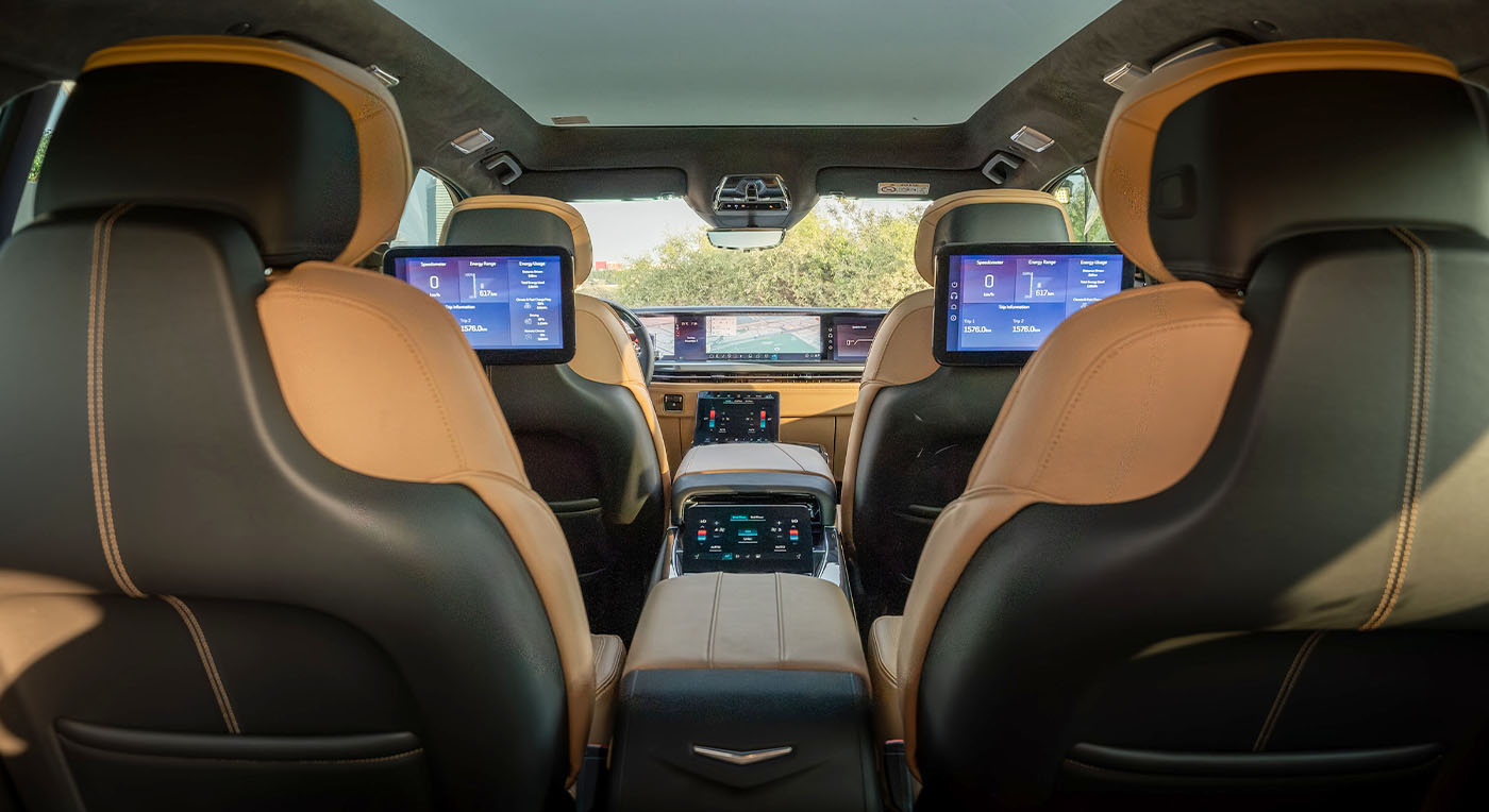 Interior of a Cadillac Escalade IQ luxury car showing the back of two front seats with sleek brown leather. Dual screens are embedded, displaying digital controls. Calm, tech-savvy ambiance.